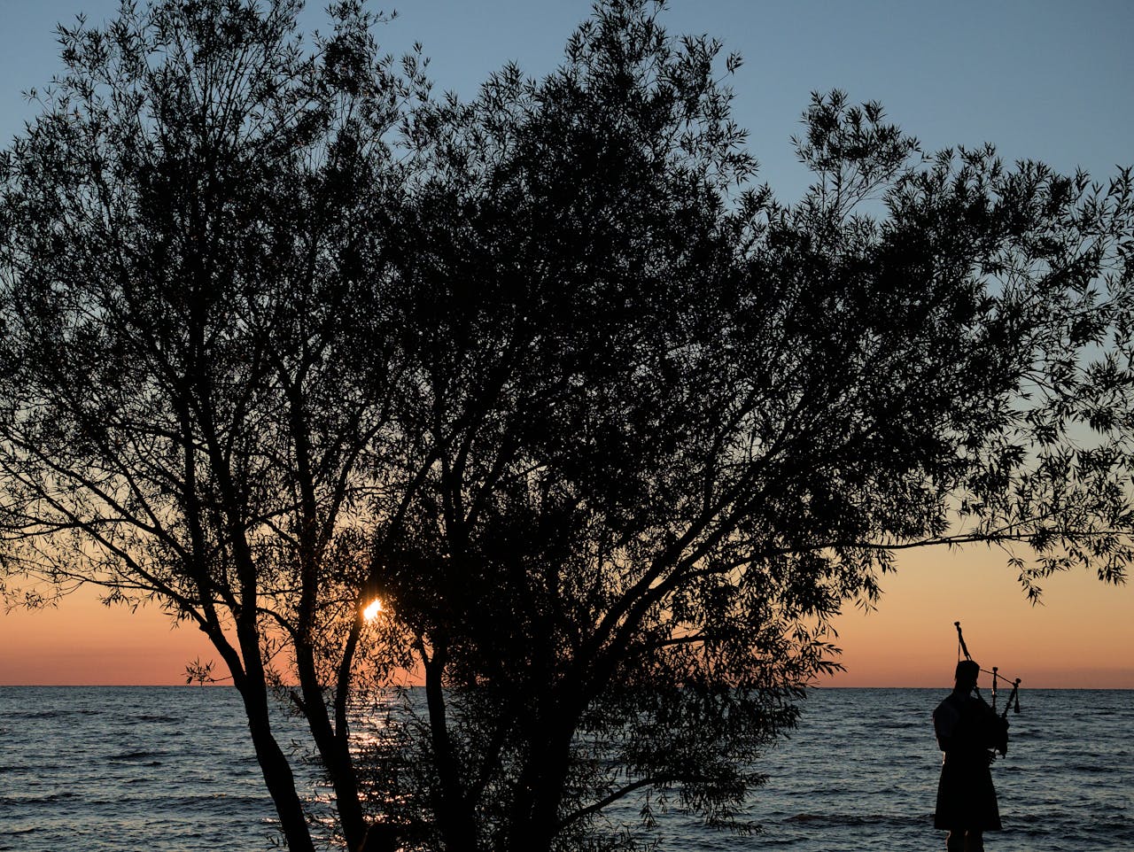 A piper silhouetted against a sunset by the ocean, framed by trees, in Southampton, Canada.